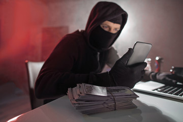 Portrait of calm hacker looking at mobile while situating at desk in apartment. Cash situating on...