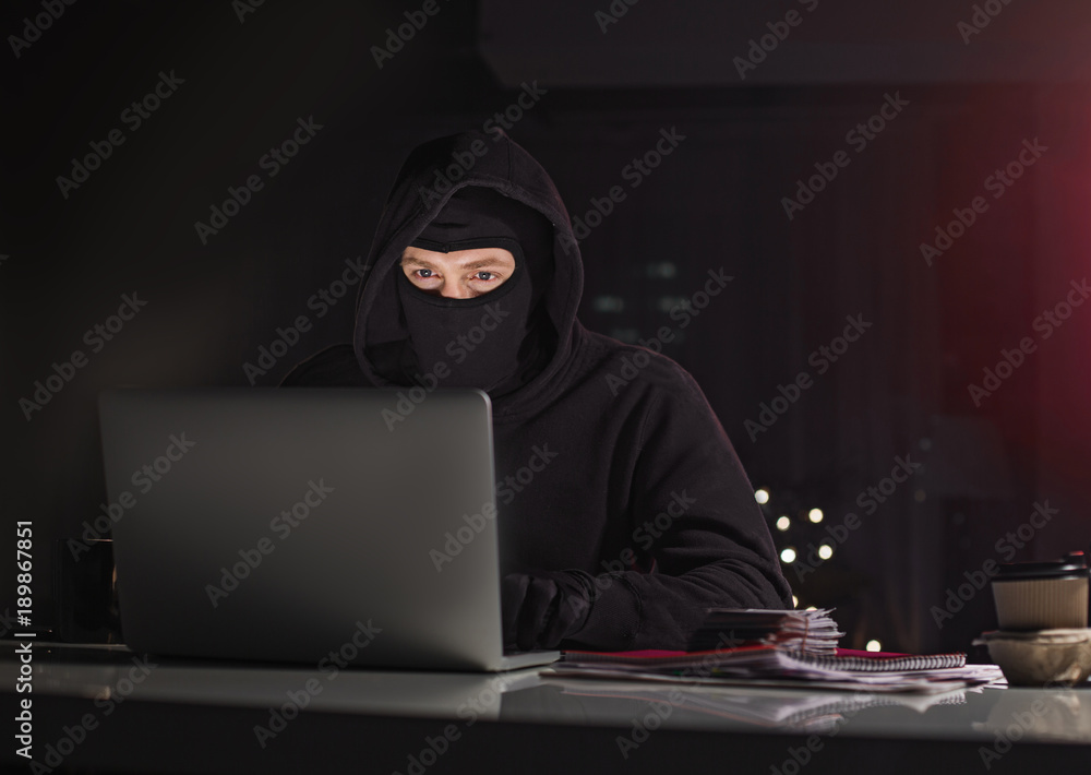 Poster portrait of orderly hacker typing in appliance while sitting at table in dark apartment. crime and d