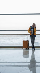 Pleasant call. Full length of cheerful young woman with suitcase is leaning on handrail at international airport and waiting for flight. She is talking on smartphone while drinking coffee. Copy space