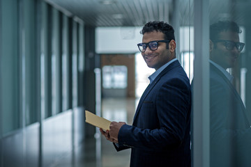 Happy glance. Joyful young elegant entrepreneur in glasses is looking at camera with smile while leaning on wall and holding documents. Copy space in the left side