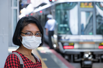 a woman with a mask on her face, standing on a platform of a railway station waiting for a train, Japan.