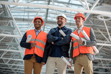 Visit object. Low angle portrait of cheerful young architects are standing in safety-helmets and looking ahead with smile while holding blueprints