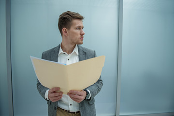 Work papers. Young man is holding documents while standing in office. He is looking aside seriously. Copy space in the right side