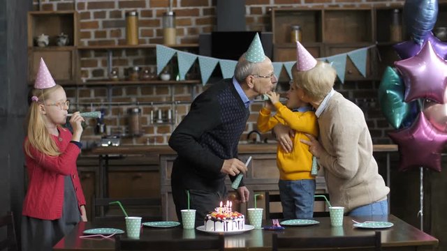 Happy multi-generation family celebrating little toddler grandson's birthday. Senior grandparents blowing horns with grandchildren and kissing their boy in decorated kitchen with baloons and cake.