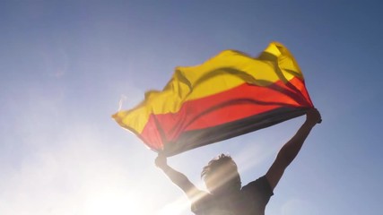 Young man holding german national flag to the sky with two hands at the beach at sunset germany - Powered by Adobe
