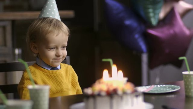 Cute smiling toddler boy with blond hair sitting at the table during his birthday party indoors. Funny child in cone hat blowing birthday horn. Cake with candles on table and baloons on background.