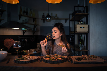 Young woman drinks wine and sits at the table with food in the modern kitchen. Romantic brunette alone dinning and dreaming in the stylish apartment in the evening. Painted heart on the wall