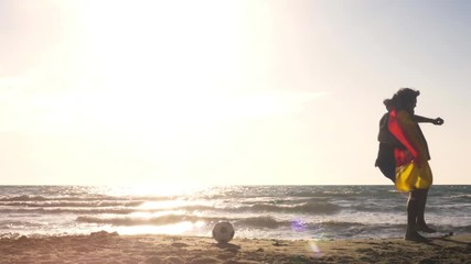 Young man in swimming trunks exulting with german flag on the sea shore at the beach at sunset silhouette with football on the sand - Powered by Adobe