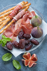 Salted breadsticks with prosciutto ham, figs and date fruits, blue stone surface, vertical shot