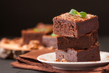 Chocolate brownie square pieces in stack on white plate with walnuts, decorated with mint leaves and cocoa on black background. Delicious dessert. Dark mood. Close up photography. Selective focus