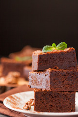 Chocolate brownie square pieces in stack on white plate with walnuts, decorated with mint leaves and cocoa on black background. Delicious dessert. Dark mood. Close up photography. Selective focus