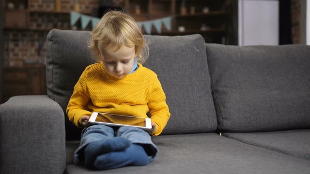 Cute concentrated little toddler boy sitting on comfortable sofa in domestic room with touchpad on his lap. Sweet blond hair boy in yellow jumper watching a cartoon on tablet pc. Dolly shot