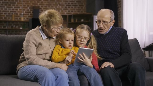 Two generations using digital touhpad together at home on the sofa. Cute blond hair grandchildren surfing the net, browsing pages and talking together with grandparents in domestic room. Dolly shot