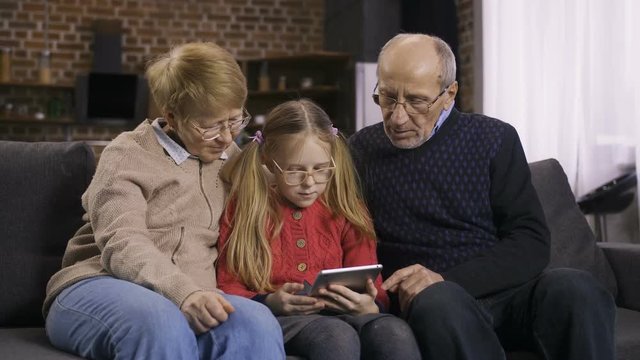 Cute blond hair teenage girl sitting on sofa together with grandparents teaching them to use touchpad. Sweet granddaughter explaining how to search and browse online on internet on tablet pc. Dolly