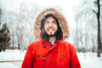 Portrait, close-up of a young stylishly dressed man smiling with a beard dressed in a red winter jacket with a hood and fur on his head. Winter and frost theme. © Elizaveta