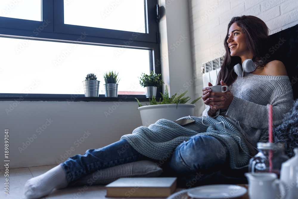 Wall mural Beautiful young woman at home drinking coffee by the window