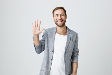 Friendly positive smiling bearded man in trendy jacket over white t-shirt and waving with his hand,...