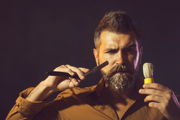 Portrait of barber in stylish clothes shows a straight razor and shaving brush. Young bearded macho...