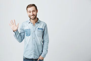 Foto op Plexiglas Friendly positive smiling young Caucasian man with stubble and dark hair in trendy denim clothes waving with hand, hailing friends while having fun indoors. Human relations and feelings © Cookie Studio