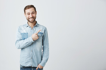 Caucasian young male in denim shirt grins at camera, indicates at copy space, advertises something....