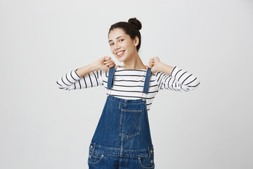 Human face expressions and emotions. Portrait of young funny positive brunette woman in sailor shirt looking at camera, having fun indoors, rejoices weekends, playing with straps of denim overall.