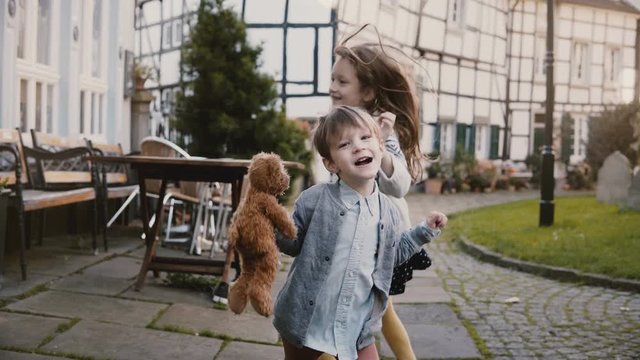 Two little kids making faces, dancing outside. Caucasian girl and boy with teddy bear. Half timber houses background 4K.