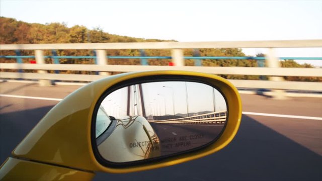 View Russian bridge (connects the mainland part of Vladivostok with Russian Island) road from the side mirror of yellow cabriolet driving along it. Early morning