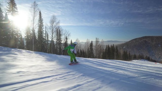 Sportsman snowboarder in bright clothes is riding down fastly from the top of a hill at winter sunny day