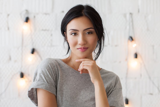 Beautiful young asian woman with brunete hair looks at camera. Cute girl posing in front of white brick wall. Smiling lady in casual dress, white wall with light bulbs on background.