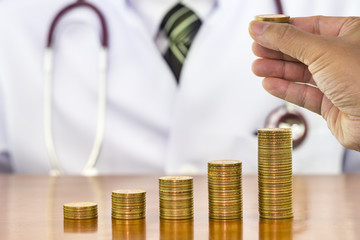 Money coins arranged as a graph with man hand holding putting on stack of money coin and blurry the doctor stethoscope around neck a scene in the back, concept of financial health and medical expenses