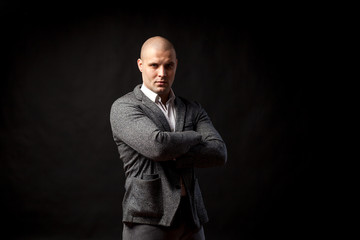 A  bald man businessman  in a white shirt, gray suit confidently looks at the camera and holds his arms crossed on his chest on a black isolated background