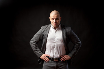 A  bald man businessman  in a white shirt, gray suit stands,  looks seriously and holds his hands at the waist on a black isolated background