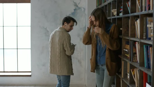 Busy male student in headphones bumping into charming blonde girl in college library. Handsome hipster helping cute female student to pick up books that fallen off and flirting with her in library.