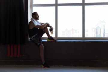Sporty active man staying near the window in the gym with bottle in hand and looking far away.