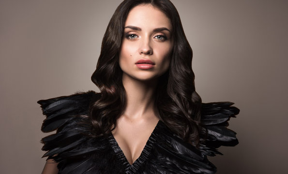 Beauty portrait of young beautiful woman in clothes with black feathers. Professional makeup, pure natural skin. Luxurious black wavy hair. Neutral brown background