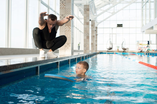 Working process in spacious swimming pool: little boy in goggles using kickboard, his middle-aged coach sitting on haunches and giving him necessary instructions while teaching him to swim