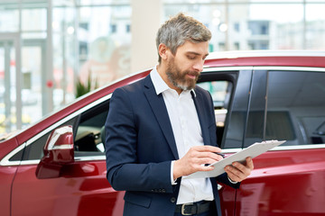 Portrait of handsome mature salesman making notes on clipboard  standing by brand new luxury car in showroom, copy space