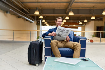 Fototapeta na wymiar Portrait of handsome young man with suitcase reading newspaper sitting in armchair while waiting for check in in modem hotel