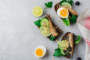 Fish sandwiches with sprats, cucumber, lime, boiled eggs, parsley leaves and mango on rye bread on a gray old concrete or stone background. Selective focus. Rustic style. Top view. Copy space.