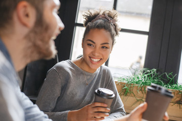 Young boyfriend and girlfriend diverse couple students having a romantic coffee-break drinking hot coffee looking at each other talking smiling close-up