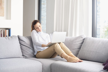 Fototapeta premium Home office. A middle aged woman making call and using her laptop while sitting on couch and working from home. 