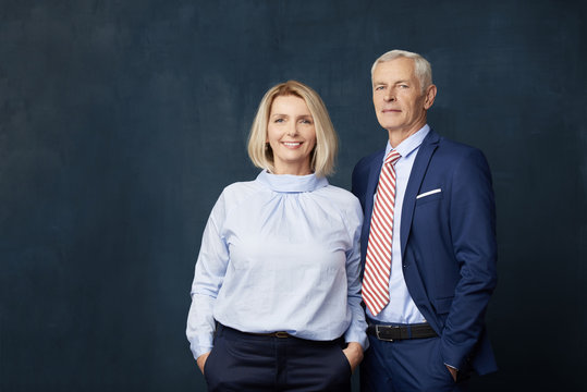 Senior couple portrait. Senior couple standing together at dark background. Beautiful blond woman looking at camera and smiling while elegant old man standing next to her. 