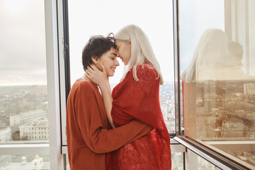 Beautiful and cute female in relationship, kissing and cuddling near open window while standing on balcony. Girlfriends bought flat together, feeling happy they started new phase in their life
