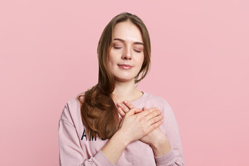 Indoor shot of younf brunette female has friendy look, keeps hands on chest, closes eyes, being satisfied with calm domestic atmopshere, isolated over pink studio background. Friendly relaxed woman