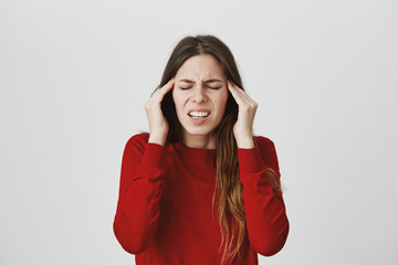 Stressed frustrated young female dressed in red sweater with closed eyes, frowns face, holds hands on temples, suffers from headache, feels annoyed and bored because of noise and useless information