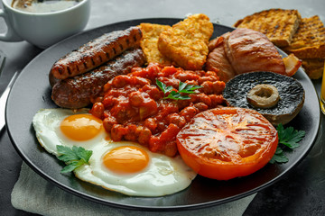 Full English breakfast with bacon, sausage, fried egg, baked beans, hash browns and mushrooms in black plate. cup coffee.