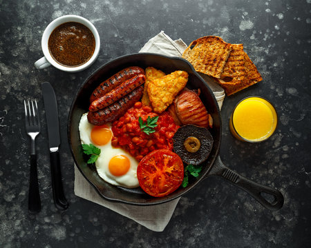 Full English breakfast with bacon, sausage, fried egg, baked beans, hash browns and mushrooms in rustic skillet, pan. cup coffe, orange juice