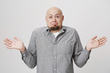 Studio shot of emotional clueless bald man dressed in gray shirt over white t-shirt having confused...