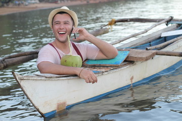 Stylish ethnic male calling by phone from a boat while drinking a fresh coconut