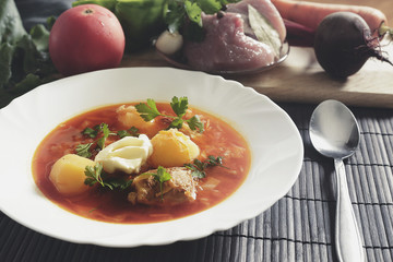 White plate with red borsch on the table near a set of fresh vegetables and meat